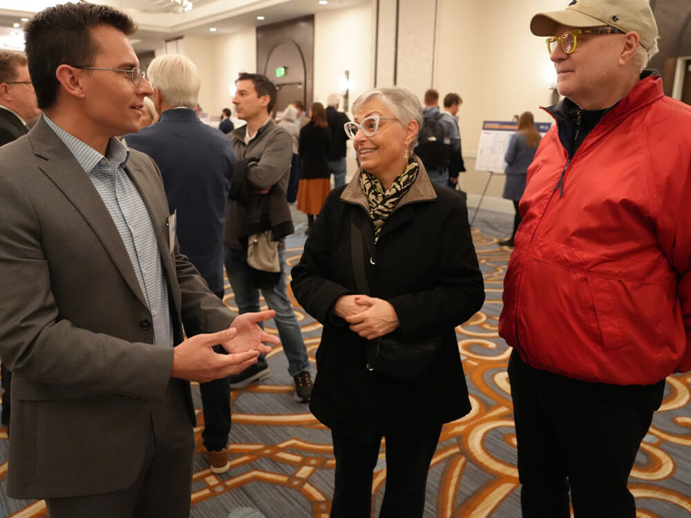 A project team member speaks with two attendees during the public information meeting. Other participants are gathered around informational boards in the background, reviewing displays and asking questions.