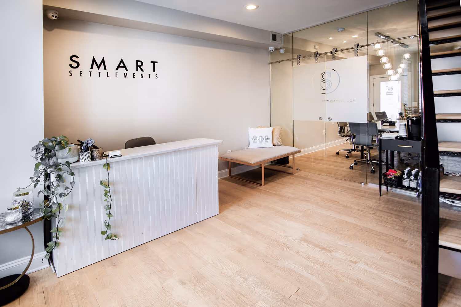 Modern office reception area with white desk, hanging plant, beige bench, glass-walled conference room, and wooden flooring.