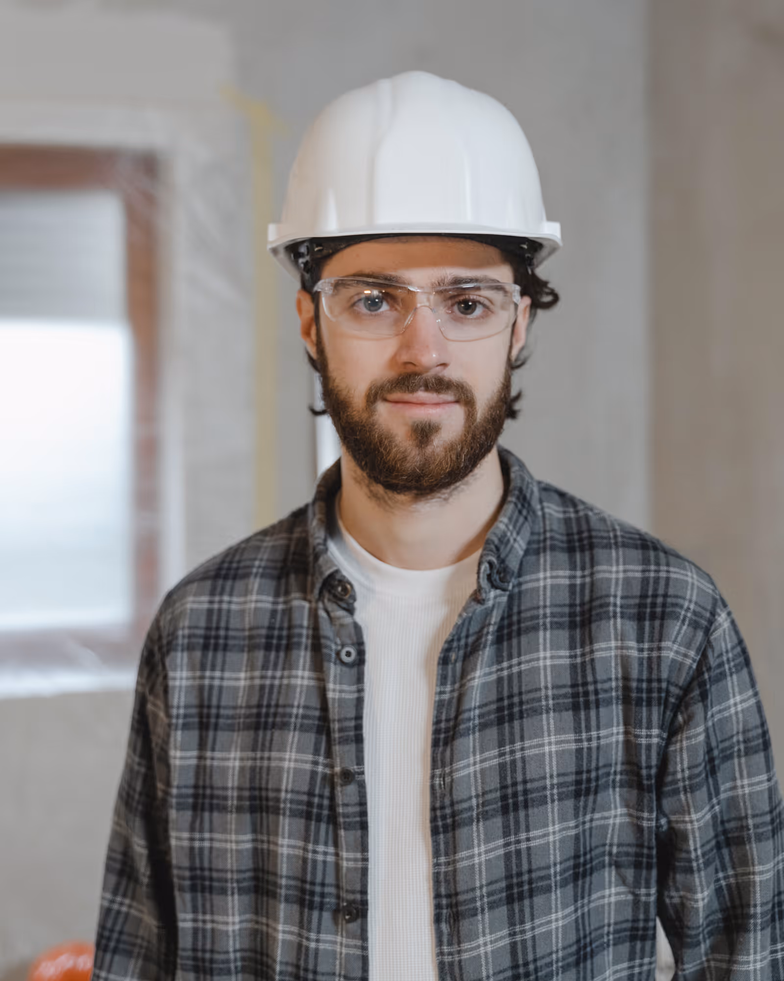 Man with beard wearing a white hard hat, safety glasses, and a gray plaid shirt standing indoors.
