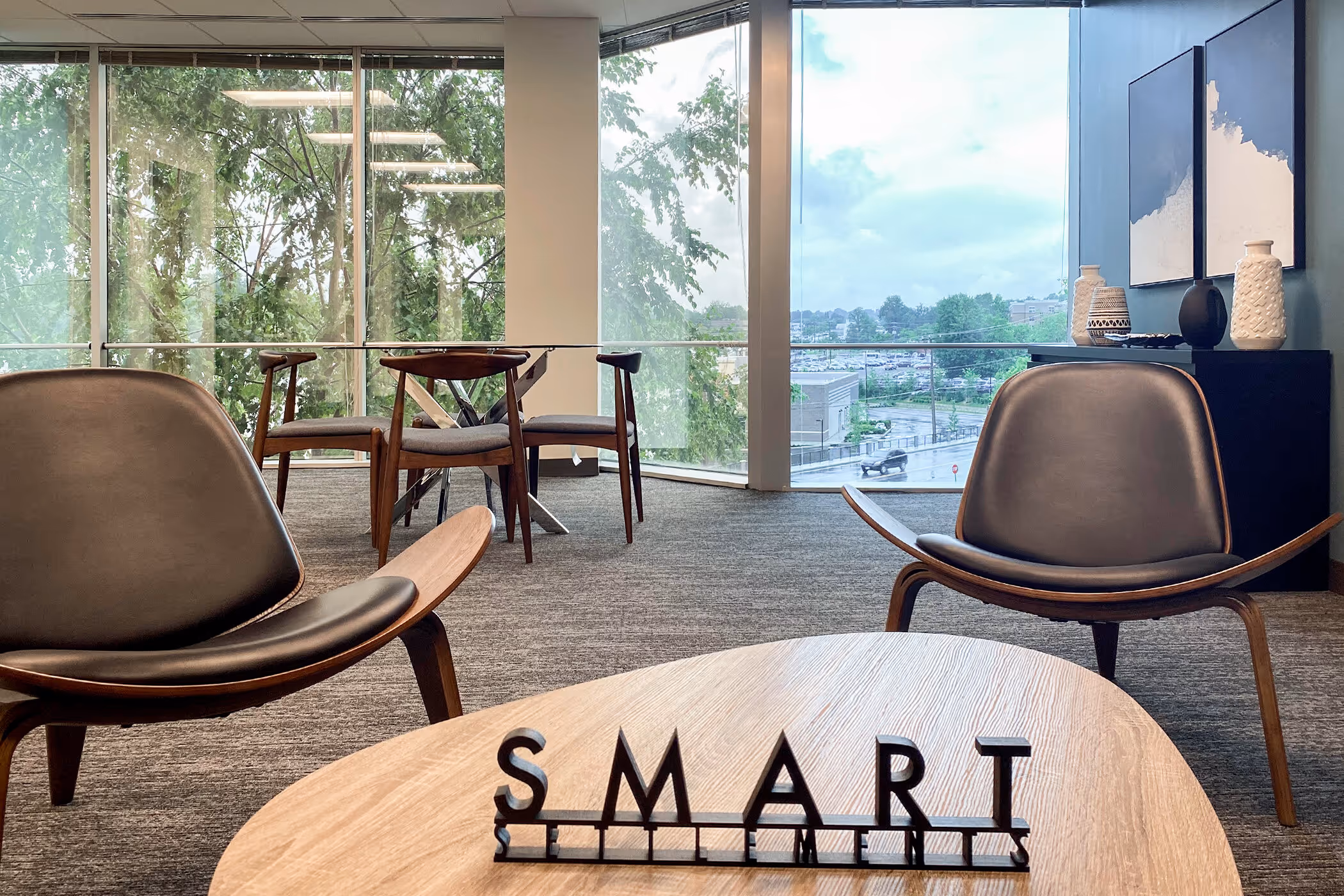 Modern office space with two mid-century style chairs, a wooden table with block letters spelling SMART, and large windows showing trees and a parking lot outside.