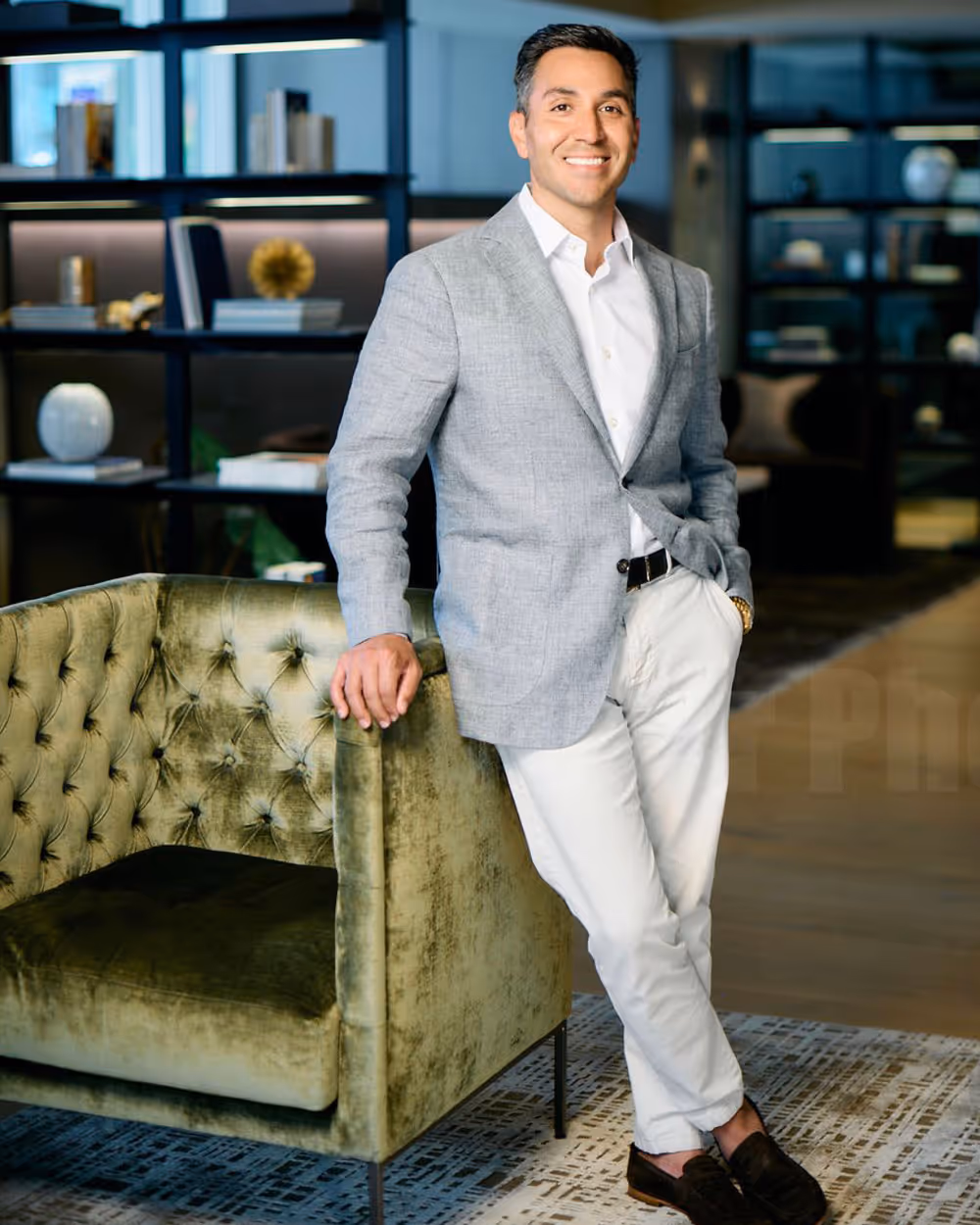 Smiling man in a light gray blazer, white shirt, and white pants leaning on a green velvet tufted armchair in a modern office setting.