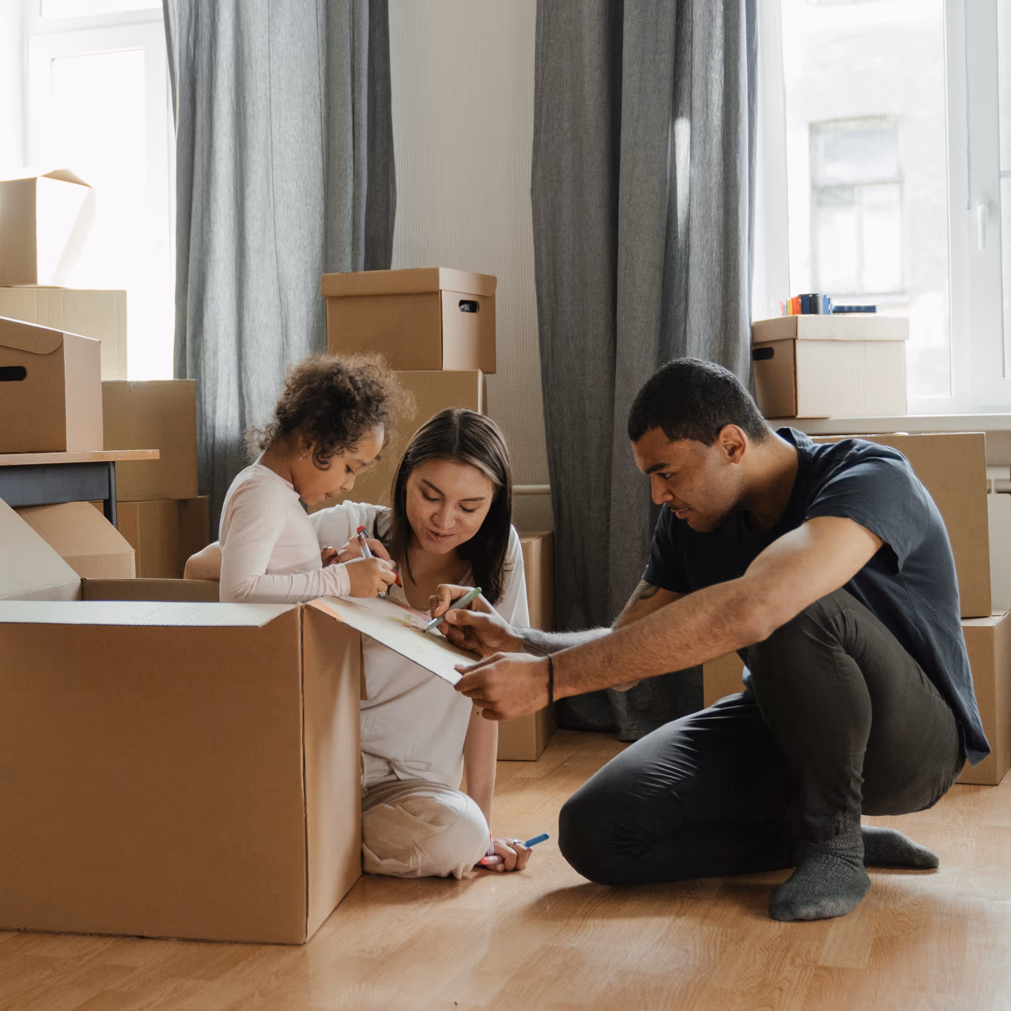 Family of three drawing on a clipboard while surrounded by cardboard moving boxes in a bright room.