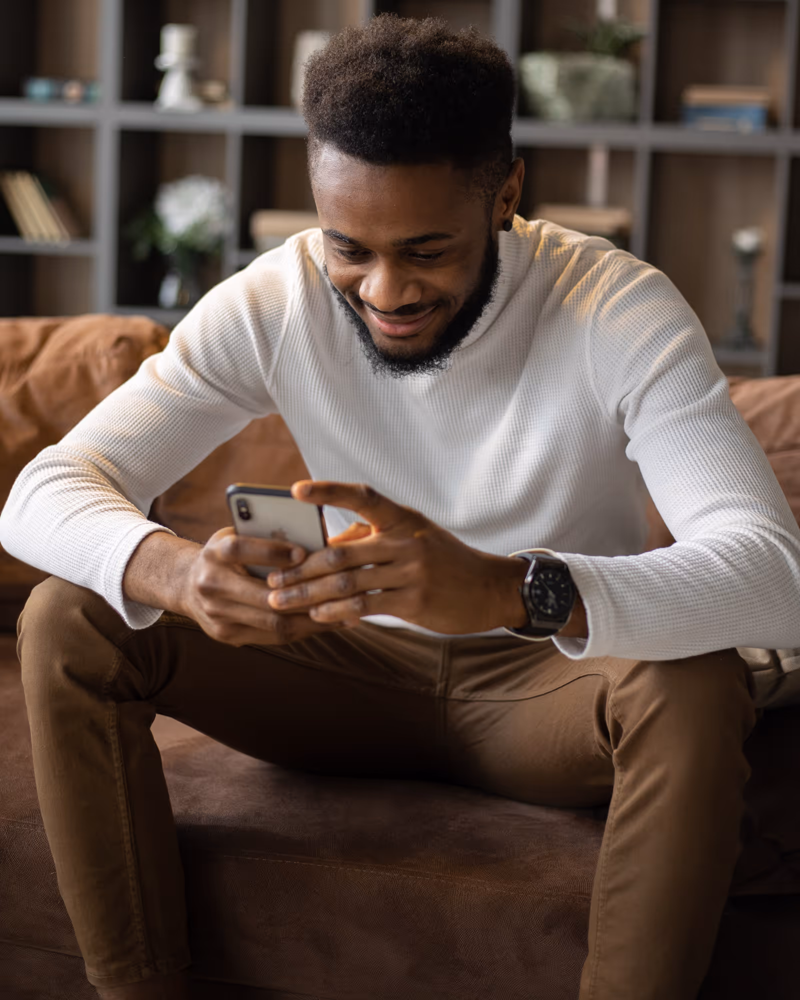 Smiling man sitting on a couch looking at his smartphone, wearing a white sweater and brown pants.