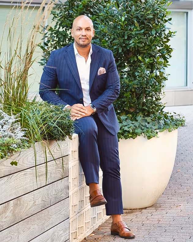 Man in a navy pinstripe suit sitting on a wooden planter box outdoors with greenery in the background.