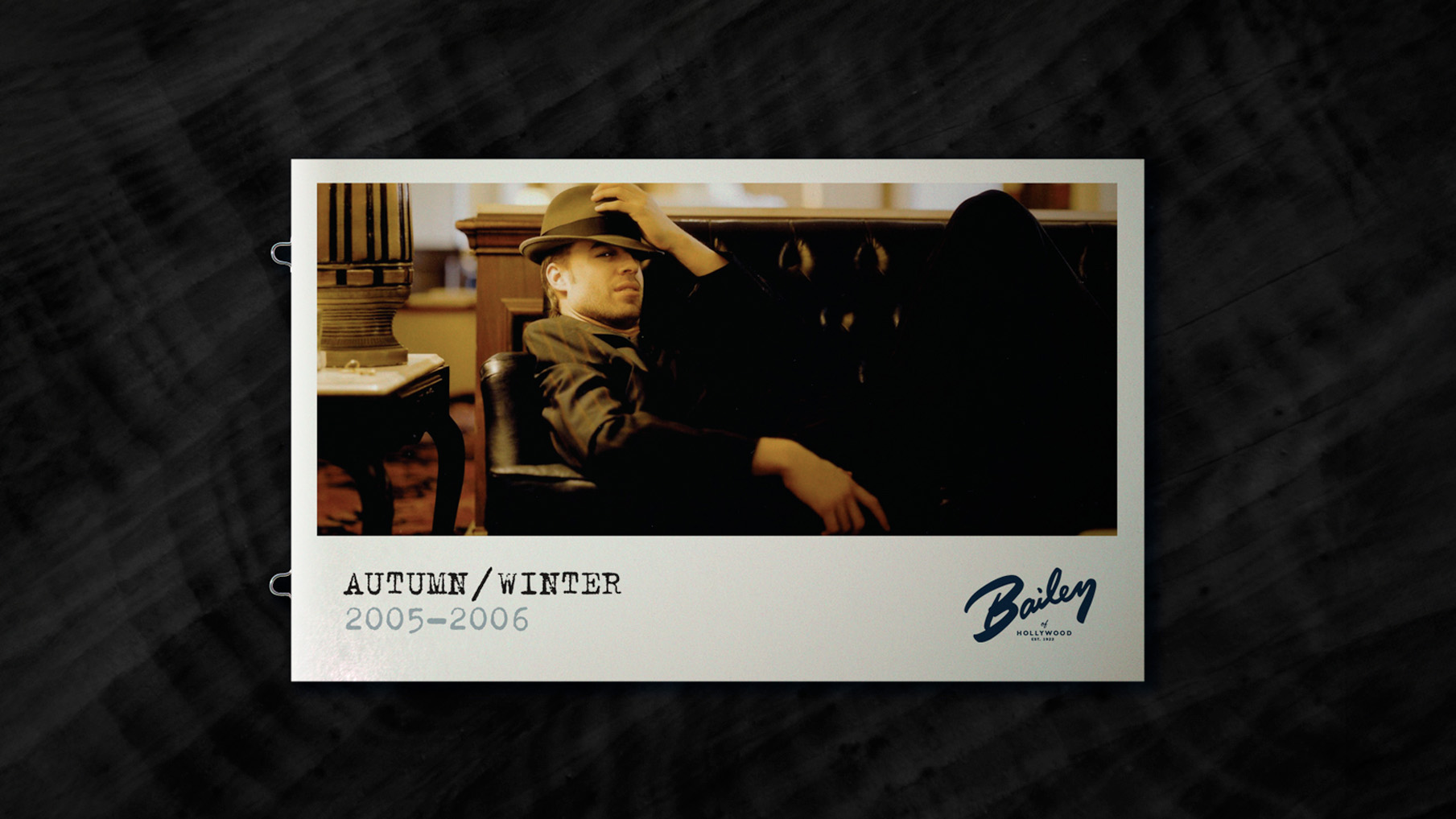 Bailey of Hollywood Autumn/Winter 2005–2006 campaign poster. A man lounges on a dark leather sofa wearing a fedora and leather jacket, tilting his hat with a relaxed, confident pose. The layout features a vintage Polaroid-style frame with the Bailey logo a
