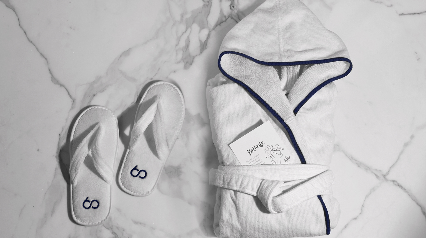 White spa-style bathrobe with navy trim neatly folded on a marble surface, accompanied by matching white slippers embroidered with the Sixty Hotels logo.