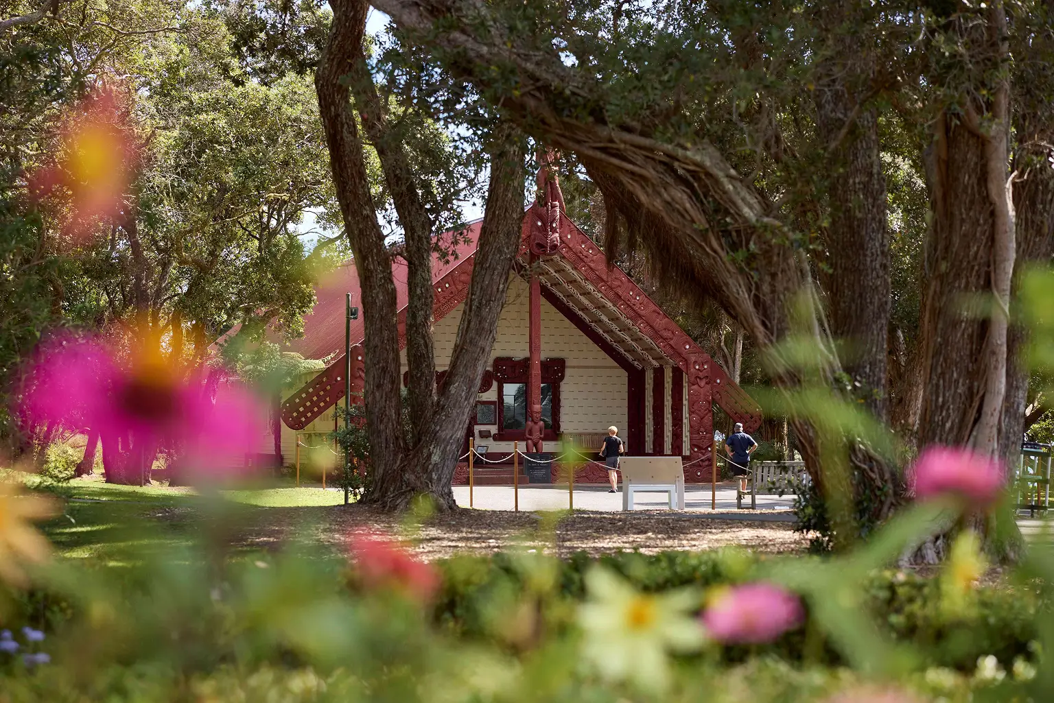 Red-roofed Maori meeting house with carvings, framed by trees and colorful flowers in the foreground.