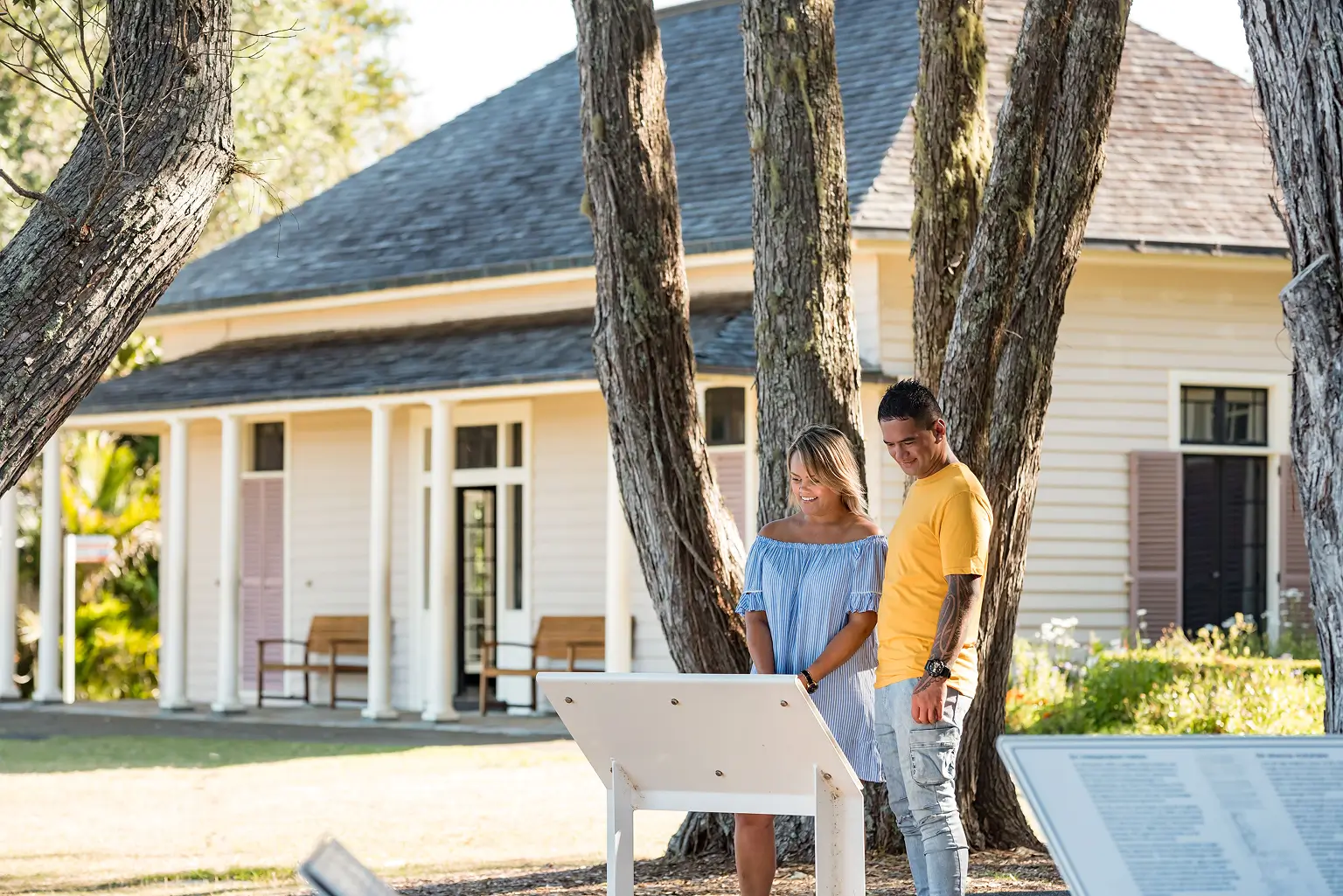 Couple reading information board outdoors near large trees and a historic white building with a porch.