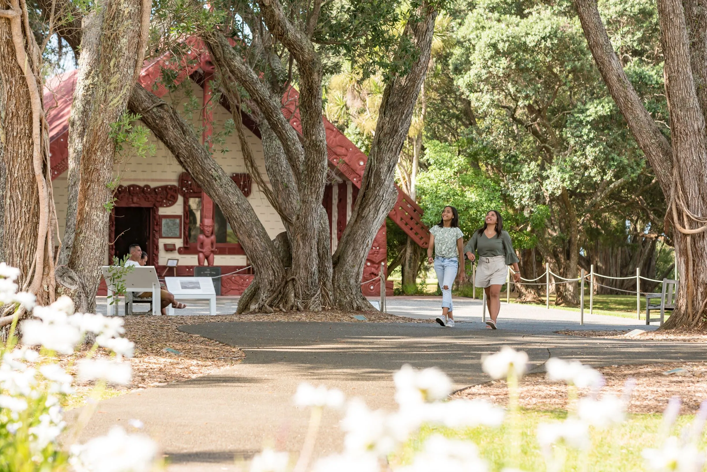 Red-roofed Maori meeting house with carvings, framed by trees and colorful flowers in the foreground.