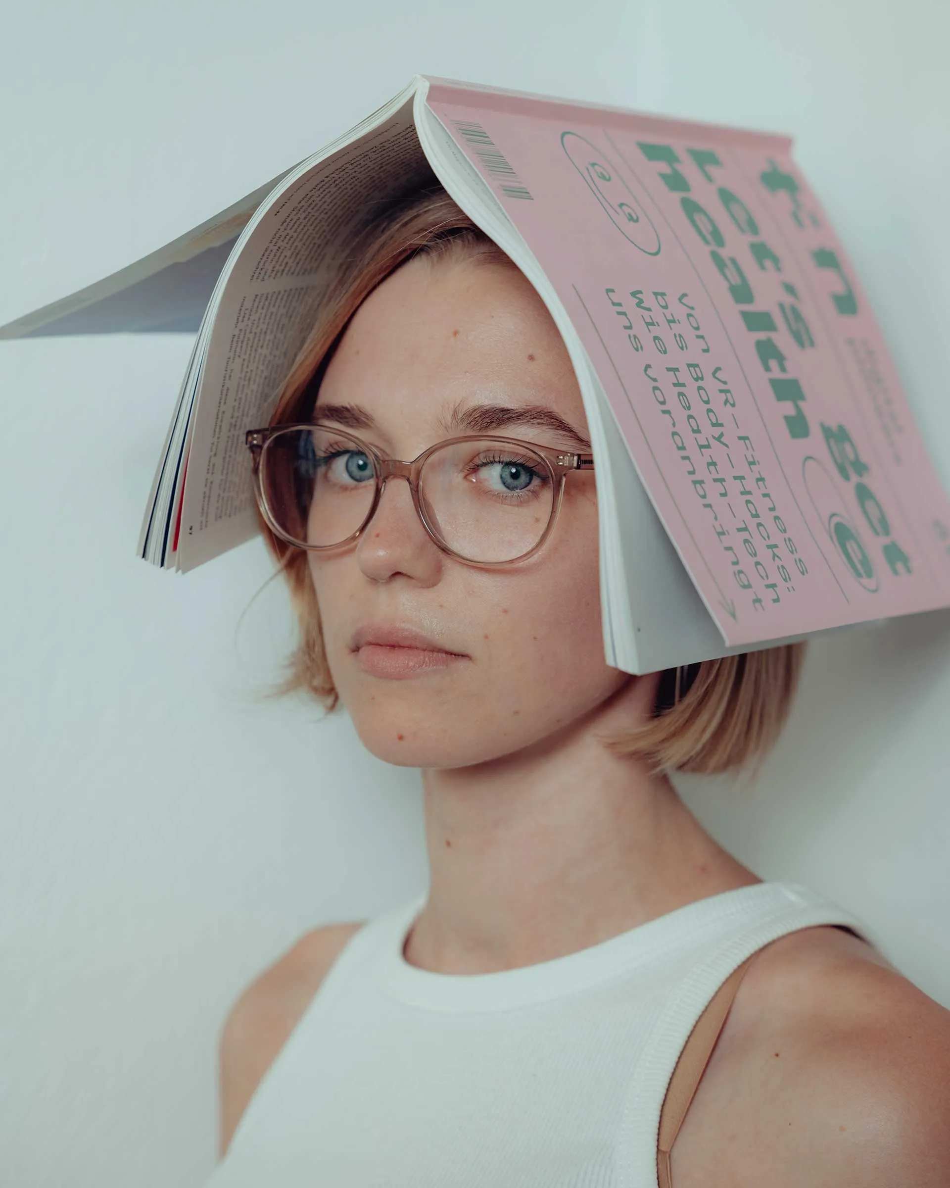 Woman with short blonde hair and glasses wearing a white tank top, holding a magazine open on her head.