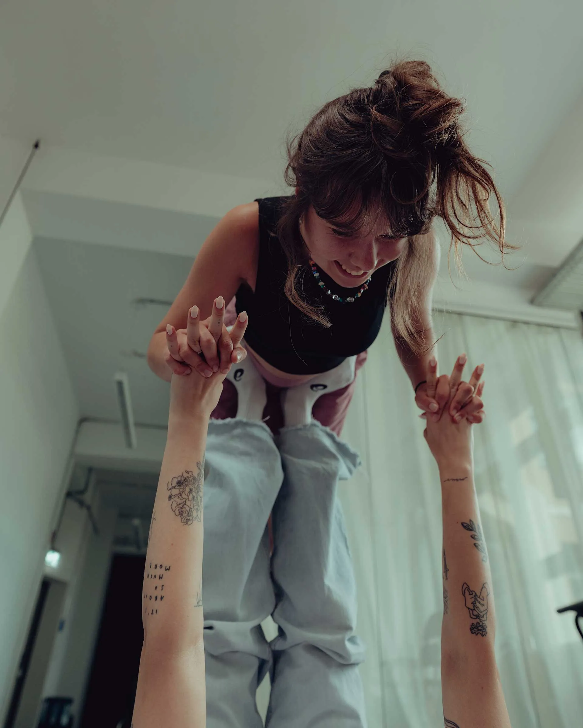 Woman with a topknot hairstyle balancing on another person's feet while holding hands indoors.