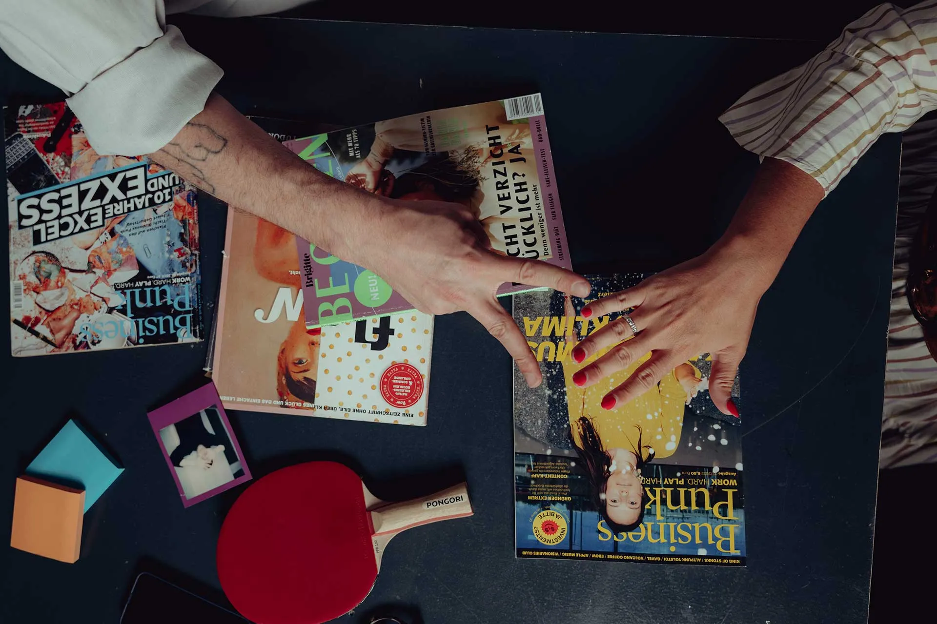 Two hands with different skin tones over magazines on a black table, accompanied by a red table tennis paddle and sticky notes.