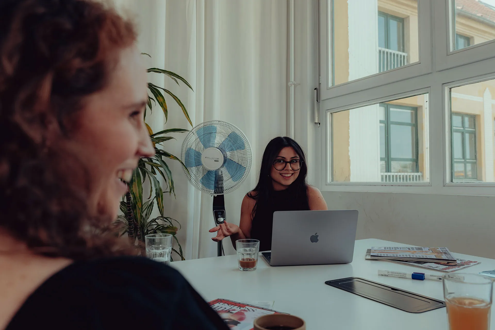 Two women in an office setting, one smiling at the camera behind a laptop and the other blurred in the foreground, with a fan and large windows in the background.