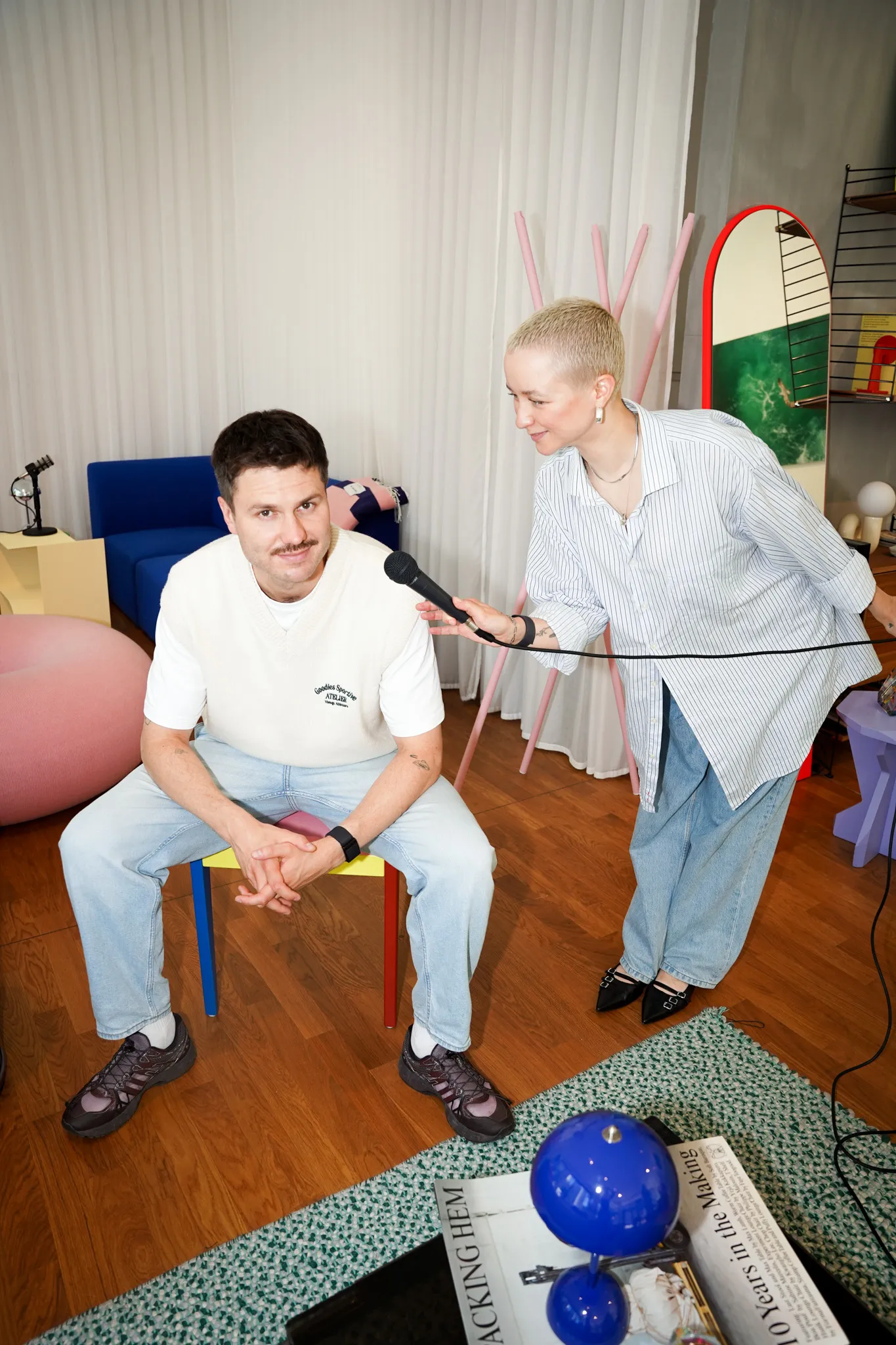 Woman holding a microphone towards a seated man in a casual modern living room with colorful furniture and wooden floor.