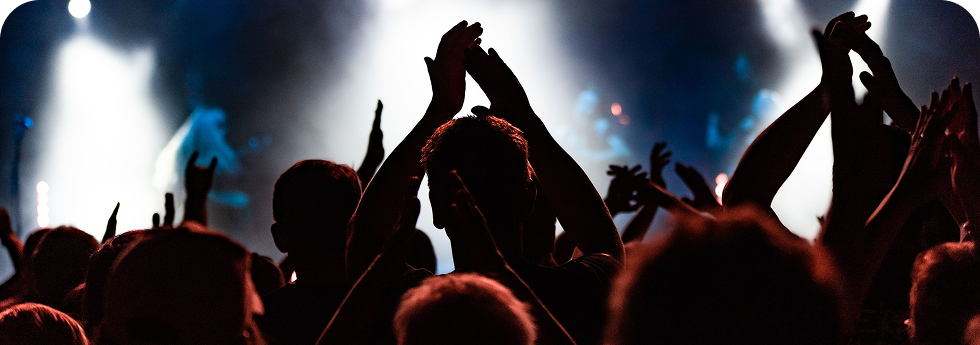 Audience silhouettes at a concert, some with raised hands and clapping, illuminated by bright stage lights and smoke effects.