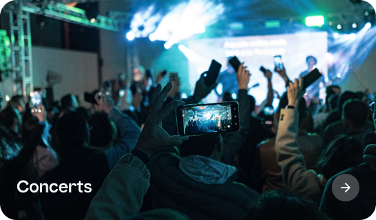 Crowd at a concert raising hands and phones to capture the stage with bright lights and performers in the background.
