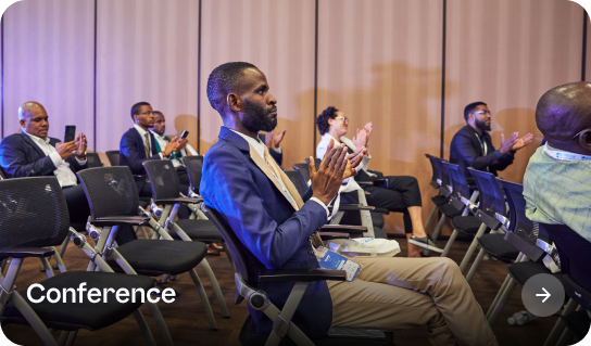 Conference attendees seated in rows of chairs, clapping, with a man in a blue blazer and beige pants in the foreground.
