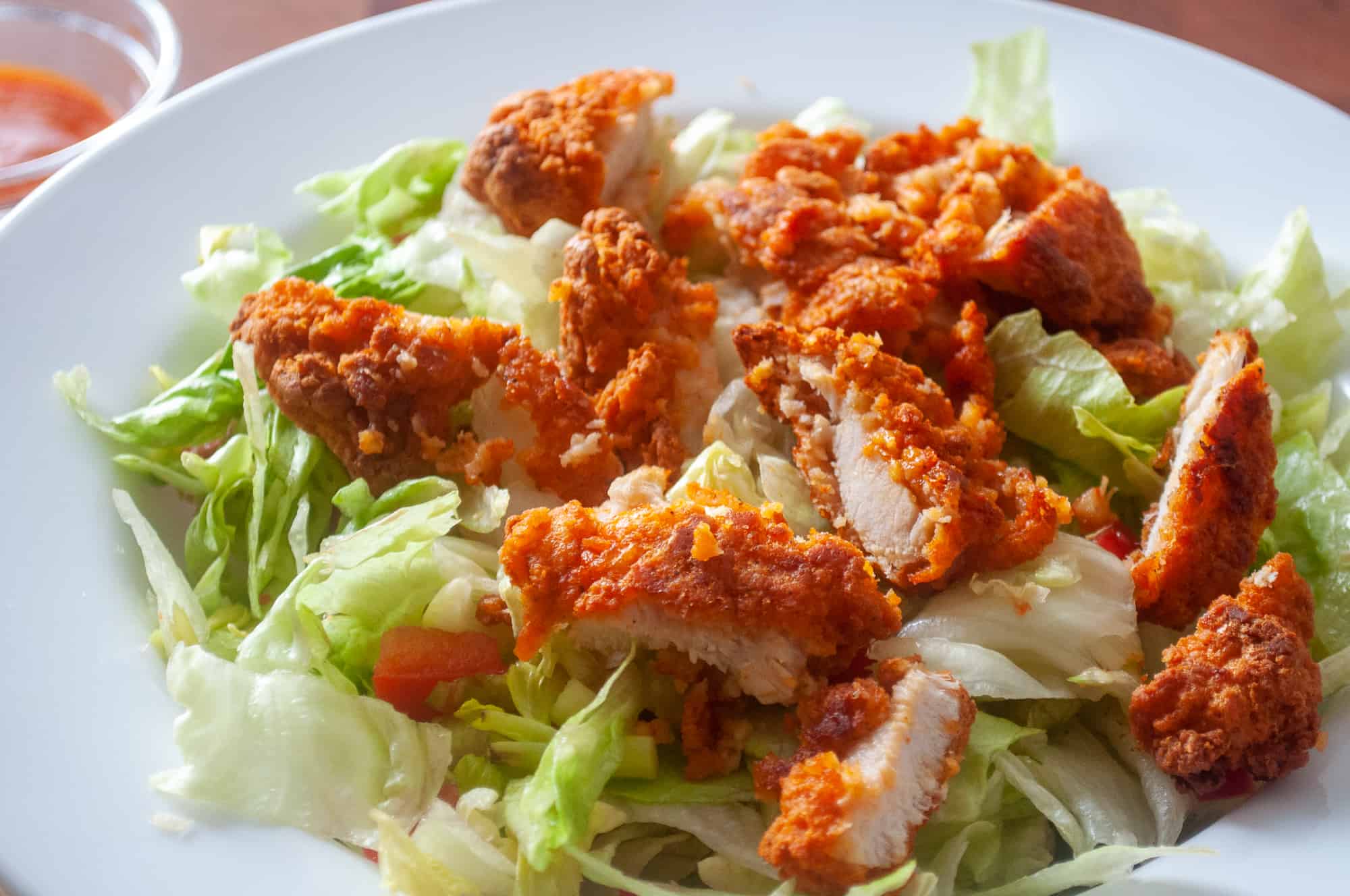 Salad with crispy fried buffalo chicken pieces and lettuce on a white plate with dipping sauce nearby.