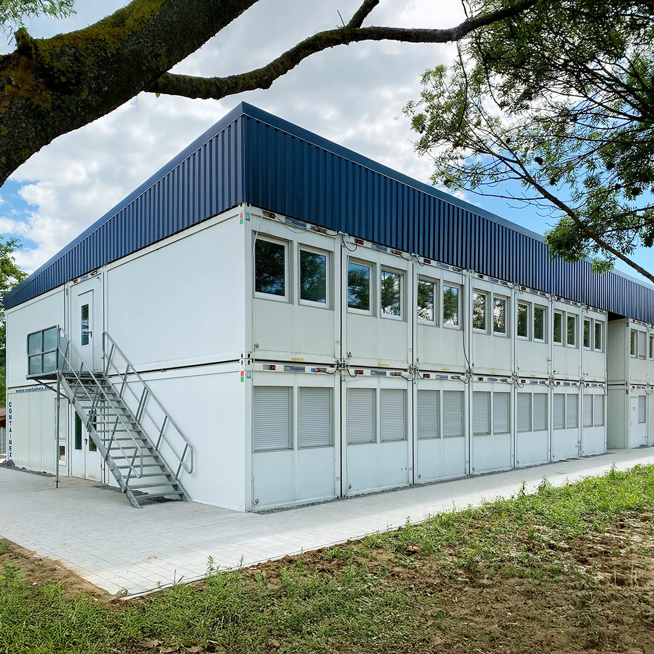 Zweistöckiges Containergebäude mit weißen Wänden, blauer Attika und Außentreppe, umgeben von Bäumen und Grünfläche.