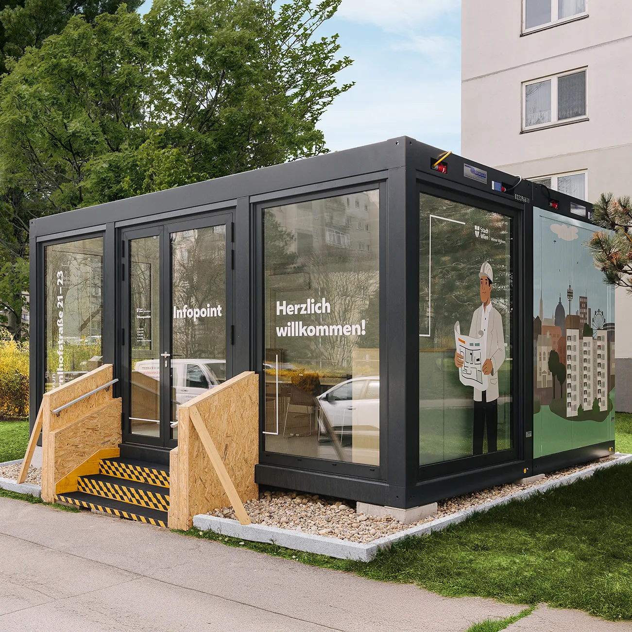 Moderner schwarzer Wohncontainer mit Infopoint und Holzgeländer an der Treppe, begrenzt von einer Wiese und einem Gehweg.