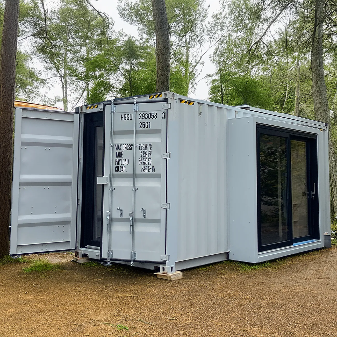 Kleines weißes Tiny House aus einem umgebauten Schiffscontainer mit geöffneten Türen und großen Fenstern in einem bewaldeten Gebiet.