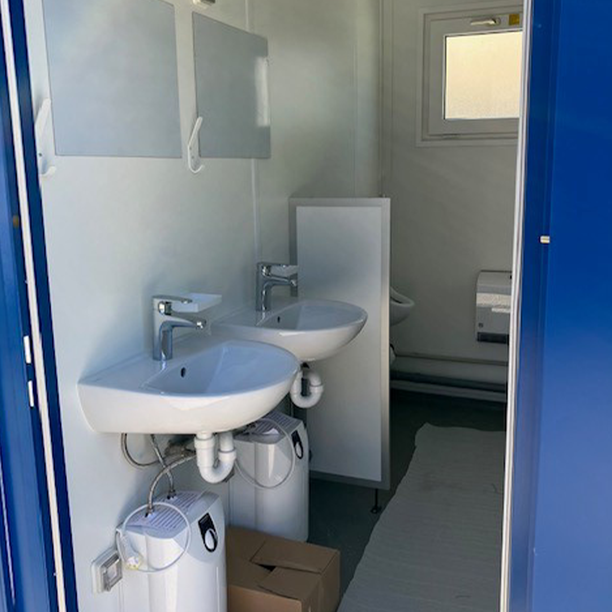 Innenansicht eines Sanitärcontainers mit zwei Waschbecken, Spiegeln, Urinal und Toilettenpapierhalter.
