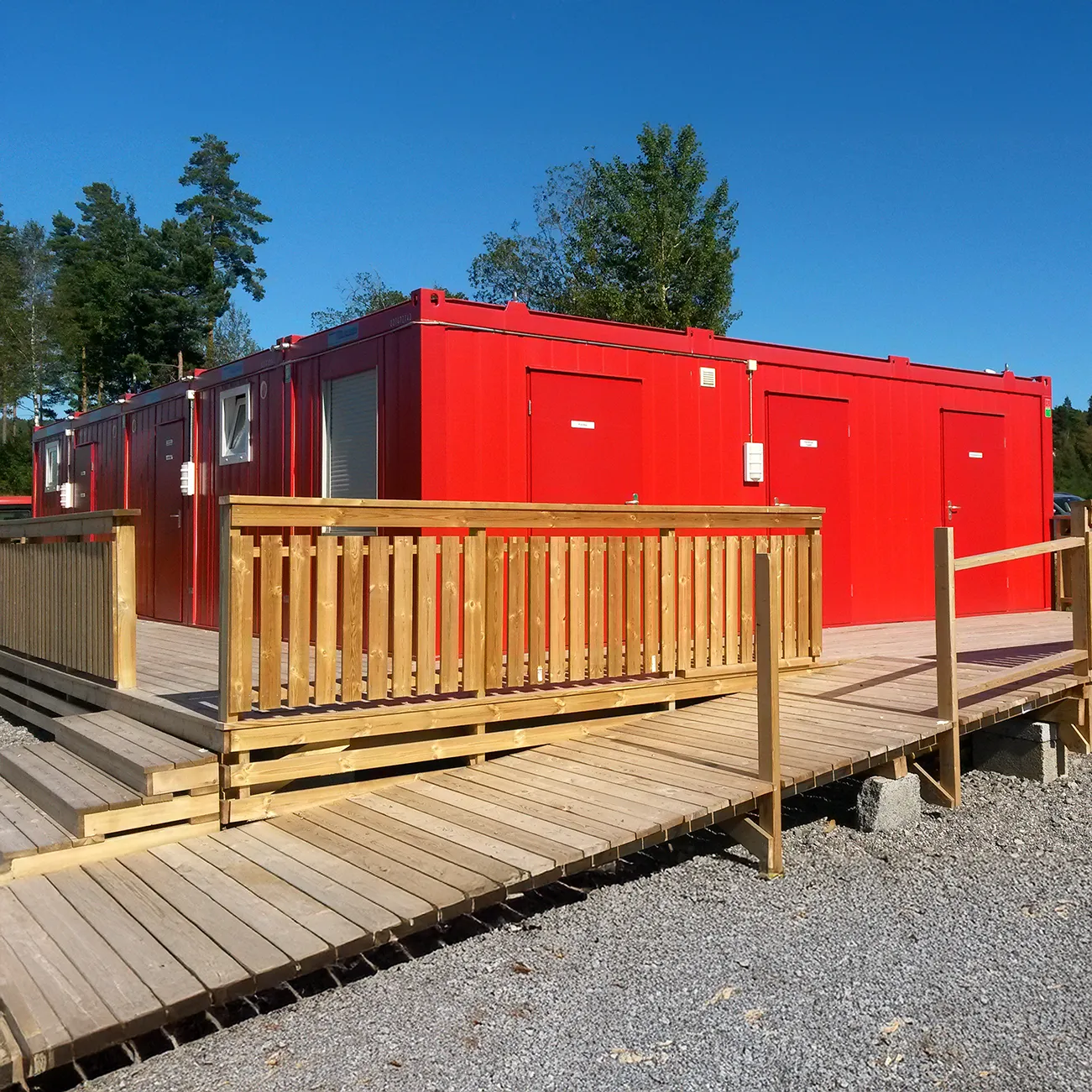 Modulare rote Sanitärcontaineranlage mit Holzterrasse und Geländer unter blauem Himmel.