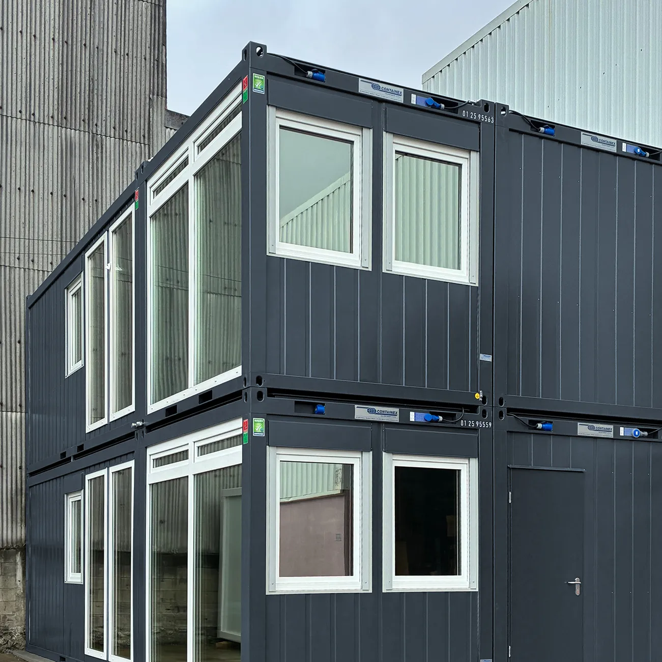 Zwei übereinander gestapelte, modulare graue Container mit weißen Fensterrahmen und einer Tür, die an einem Industriegelände stehen.