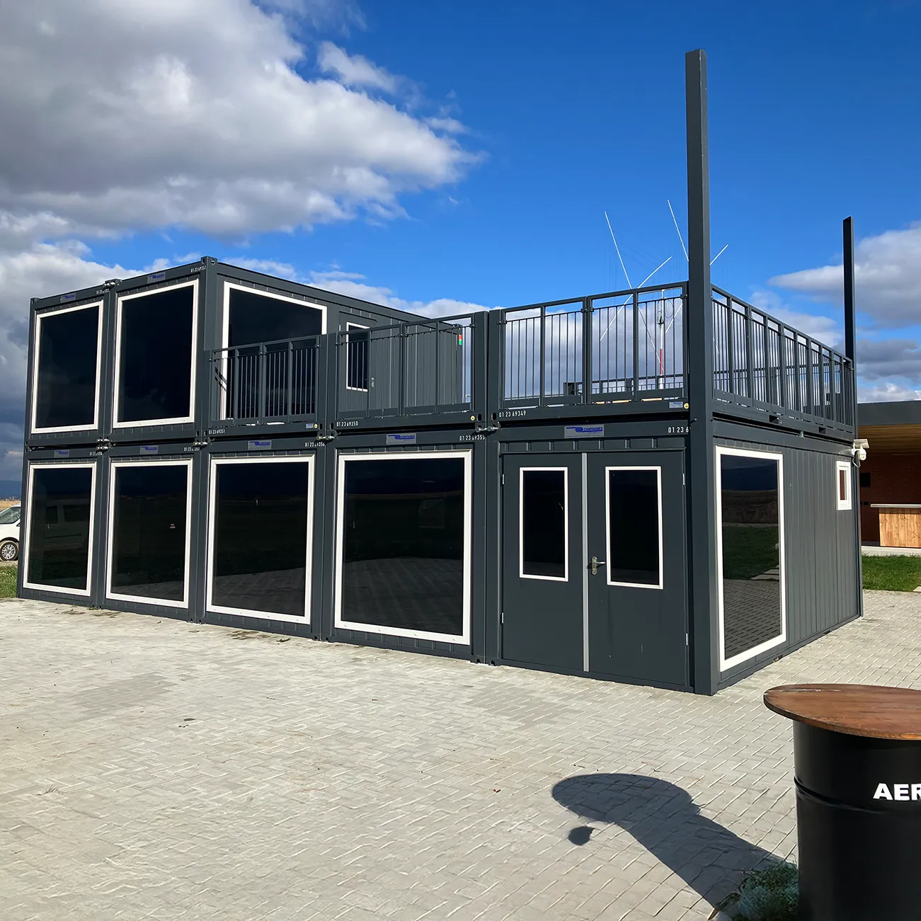 Modulare Containeranlage mit zwei Stockwerken, schwarzen Wänden und großen Fenstern unter einem blauen Himmel mit Wolken.