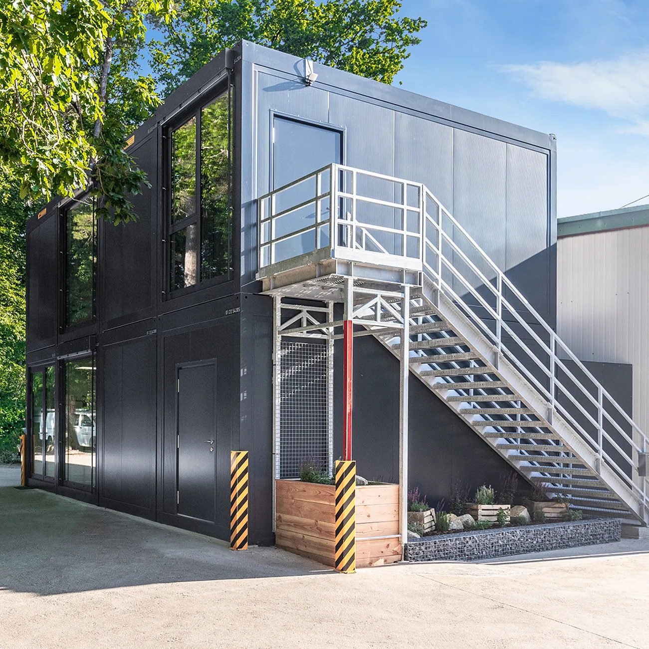 Modulare zweistöckige Containeranlage mit Außentreppe und großen Fenstern, umgeben von Bäumen.