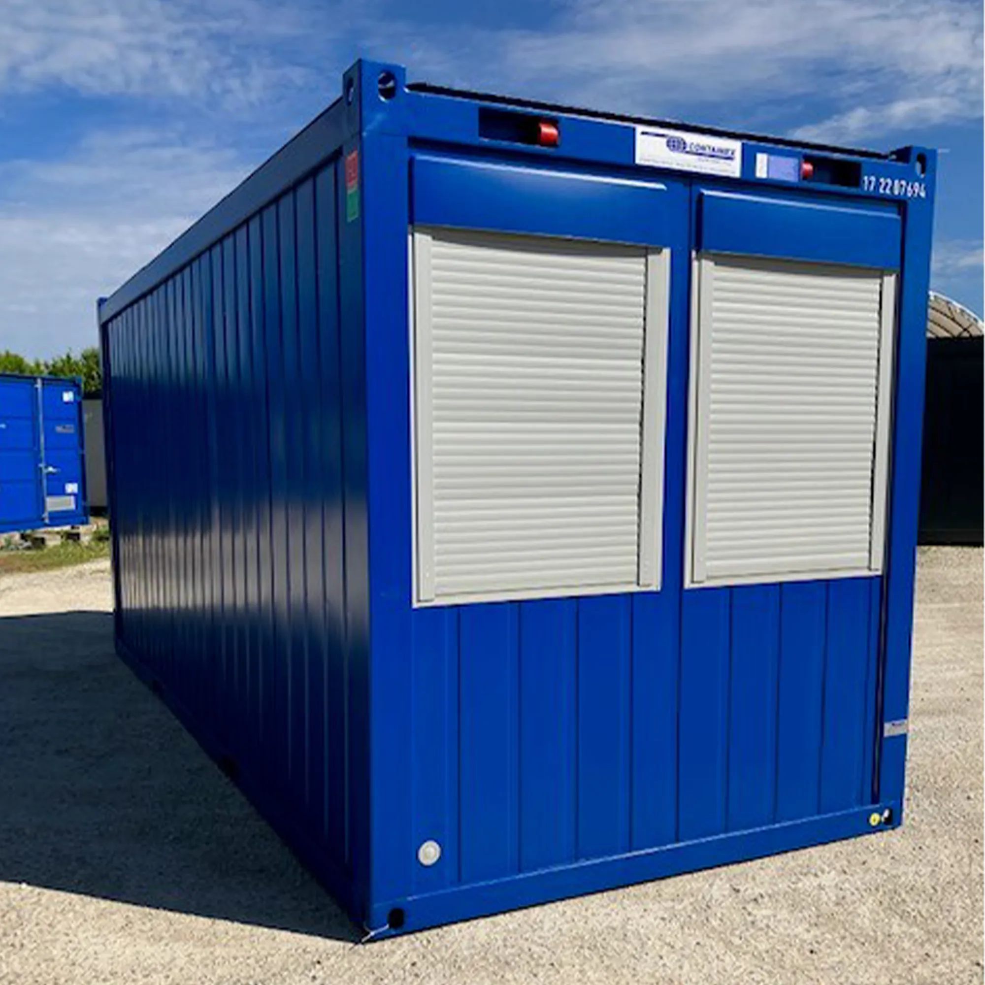 Blauer Bürocontainer mit zwei geschlossenen weißen Rollläden, steht auf geschottertem Boden unter bewölktem Himmel.