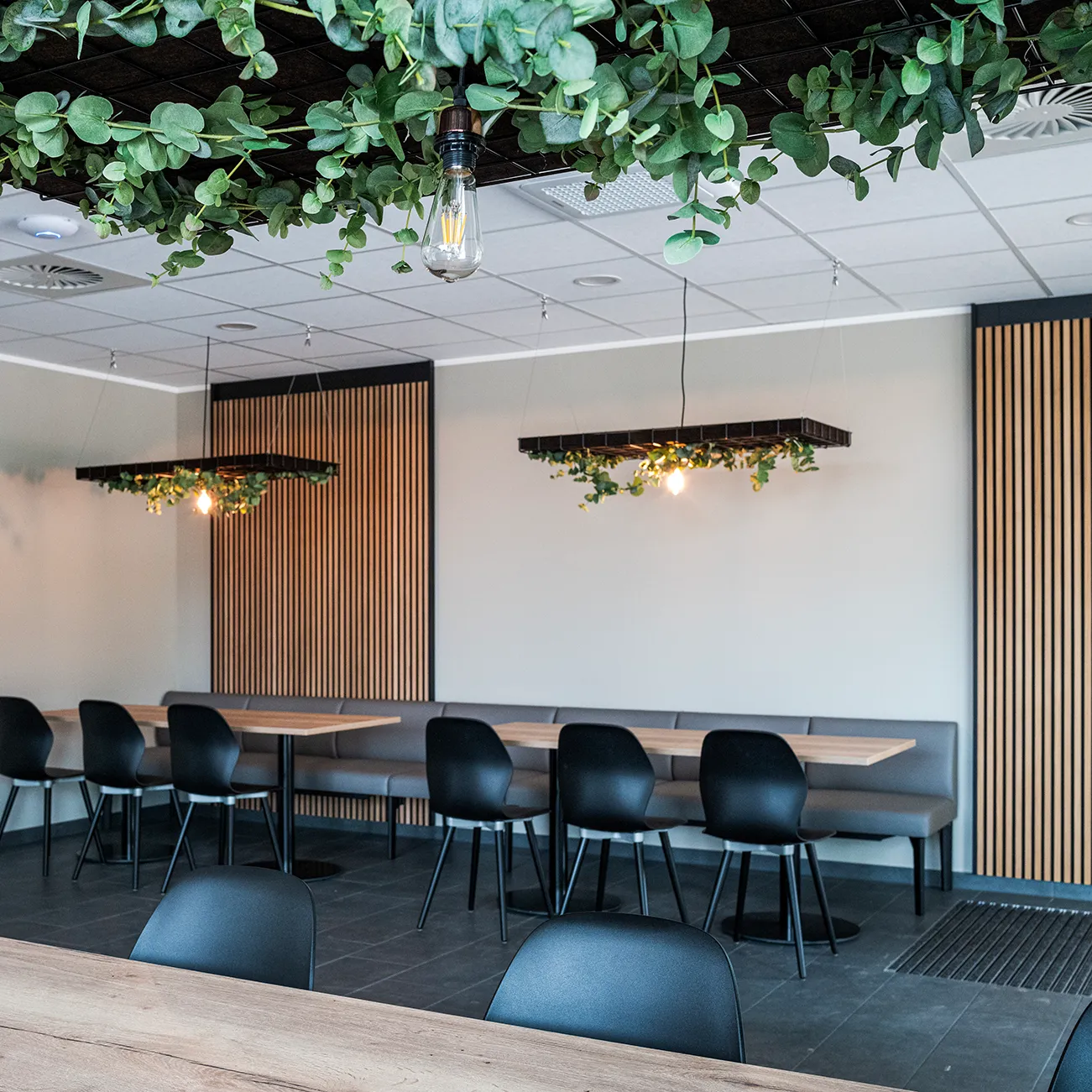 Moderner Besprechungsraum mit schwarzen Stühlen, Holztischen und langen grauen Sitzbänken, Deckenlampen mit grüner Begrünung und warmem Licht, helle Wände mit vertikalen Holzpaneelen.