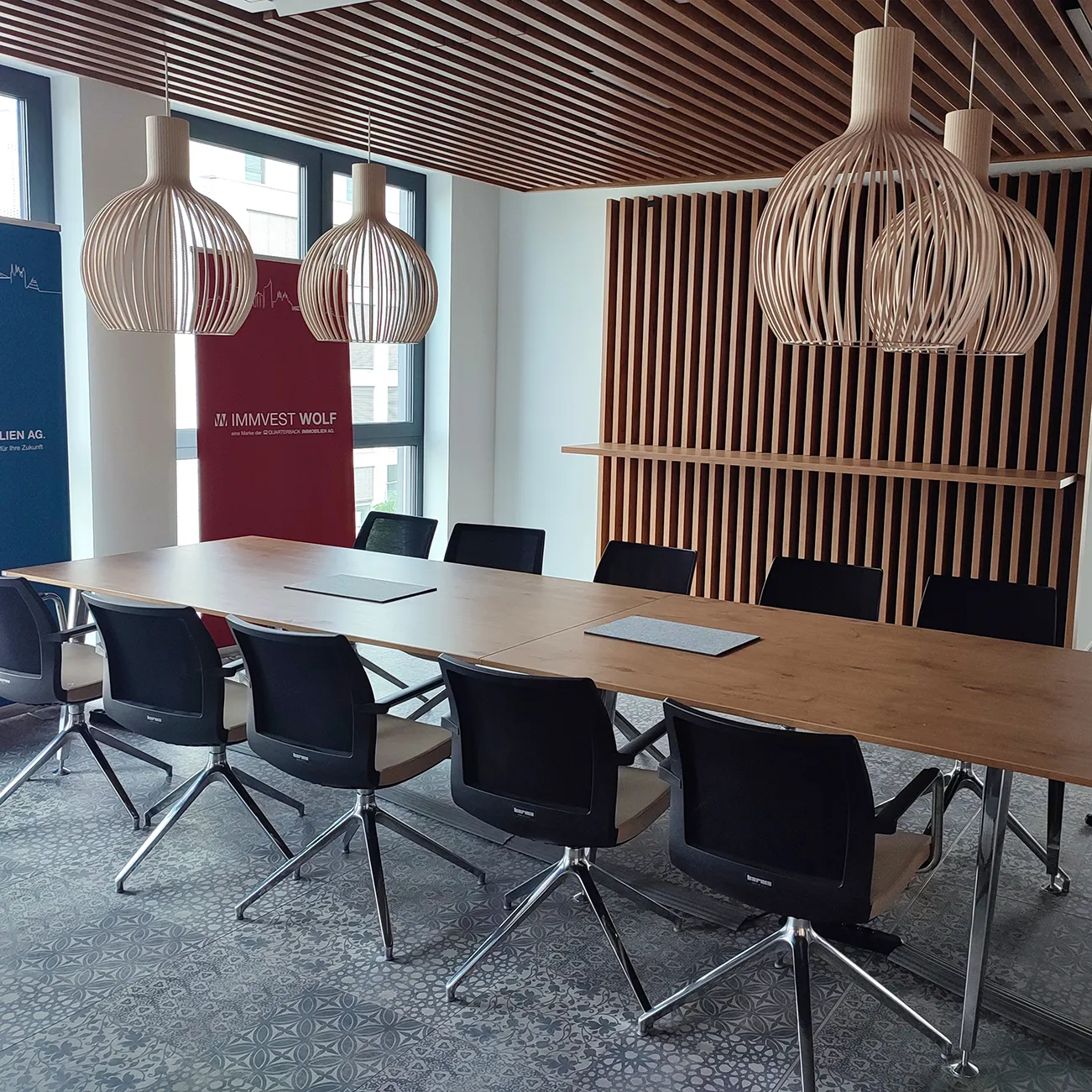 Moderner Besprechungsraum mit langem Holztisch und zehn schwarzen Drehstühlen, hellen, künstlerischen Holzpendelleuchten und einer Holzlamellenwand mit eingebautem Regal.