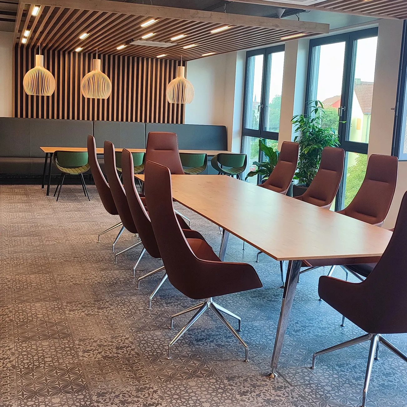 Moderner Meetingraum mit großem Holzkonferenztisch, acht dunkelrote Drehstühlen, bodentiefen Fenstern und grünen Pflanzen sowie einer Sitzecke mit grünen Stühlen und Holzlamellen an Wand und Decke.