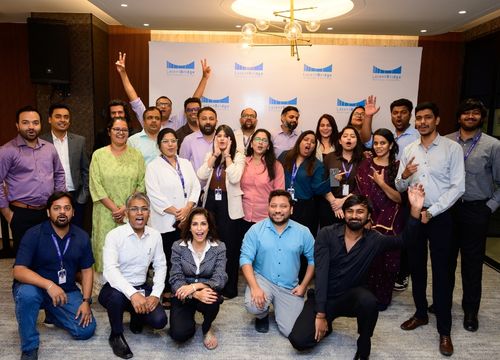 Group of eight adults smiling and posing for a selfie in a warmly lit indoor venue with tables and chairs in the background.
