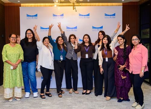 Group of eight adults smiling and posing for a selfie in a warmly lit indoor venue with tables and chairs in the background.