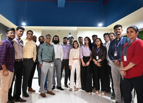 Group of eight adults smiling and posing for a selfie in a warmly lit indoor venue with tables and chairs in the background.