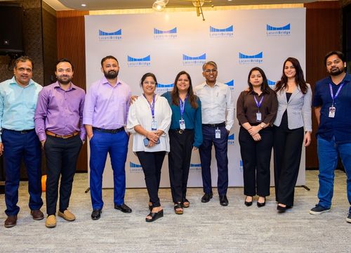 Group of eight adults smiling and posing for a selfie in a warmly lit indoor venue with tables and chairs in the background.