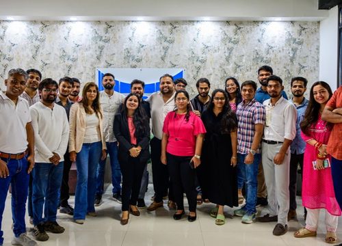 Group of diverse colleagues seated around a conference table with drinks, notebooks, and pastries, engaged in a meeting in a modern office with large windows showing city buildings.