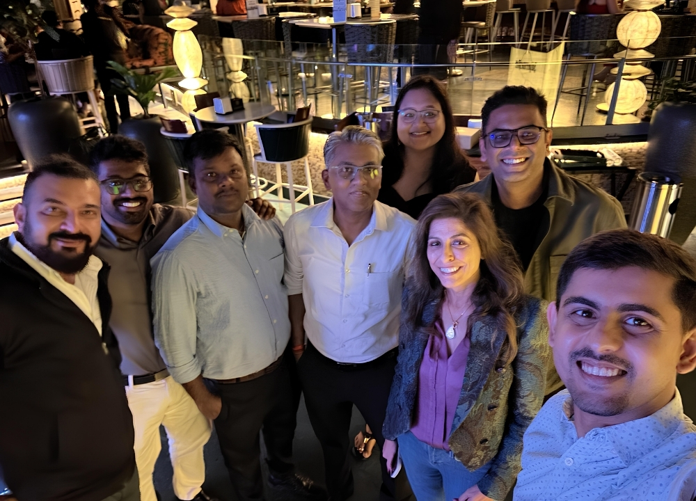 Group of eight adults smiling and posing for a selfie in a warmly lit indoor venue with tables and chairs in the background.