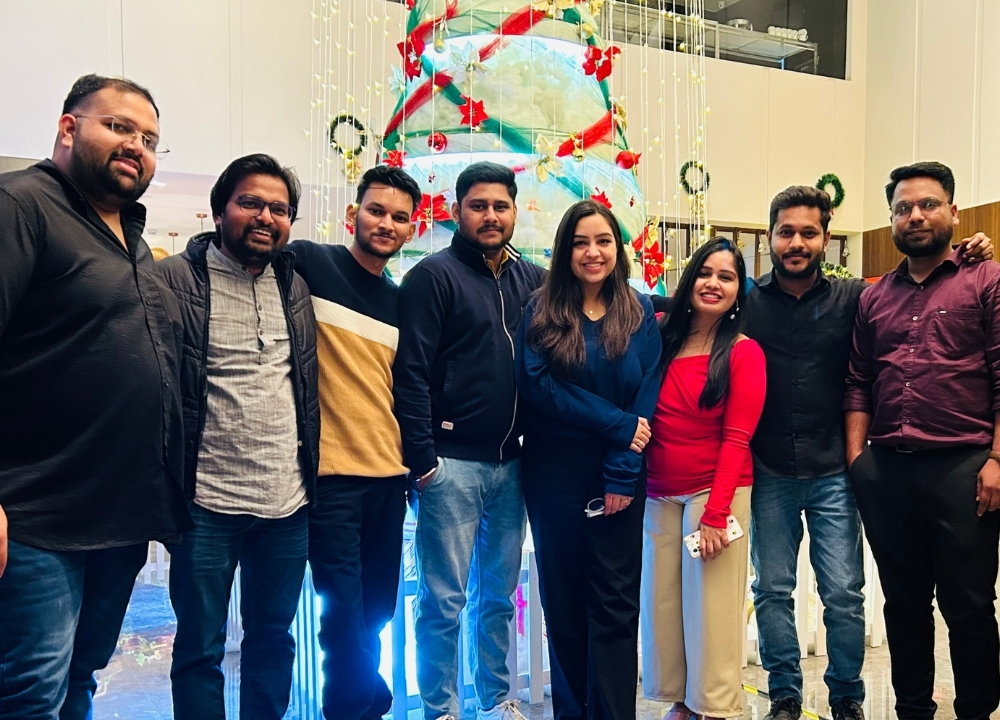 Group of eight adults smiling and posing for a selfie in a warmly lit indoor venue with tables and chairs in the background.