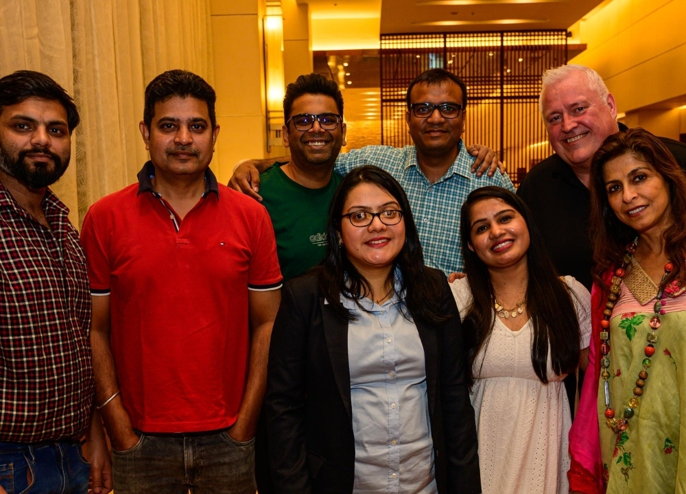 Group of eight adults smiling and posing for a selfie in a warmly lit indoor venue with tables and chairs in the background.
