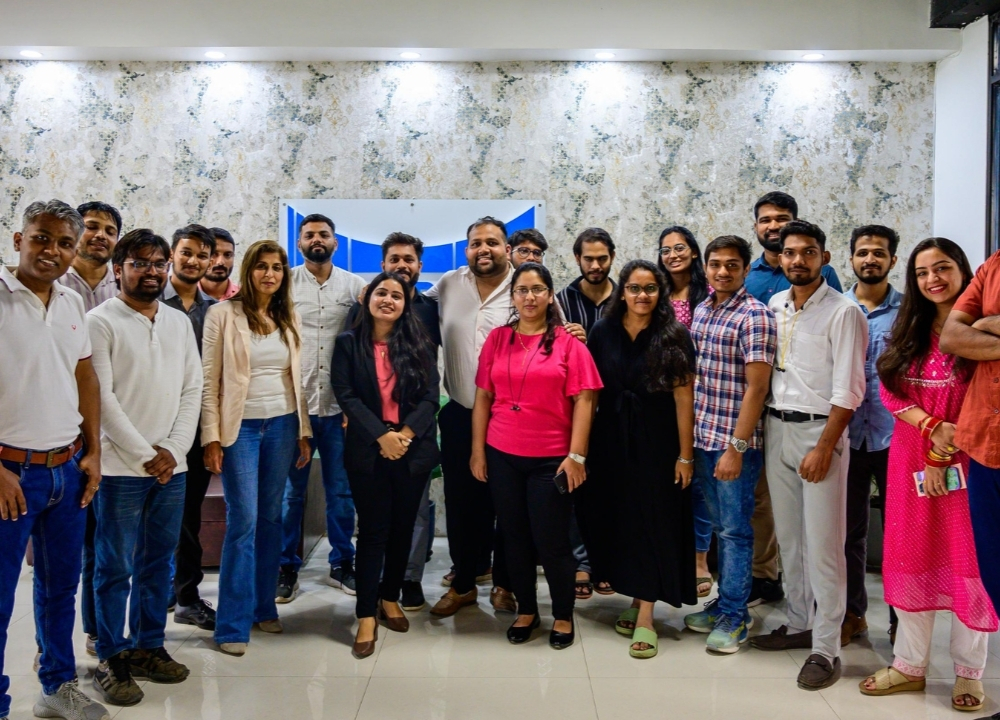 Group of eight adults smiling and posing for a selfie in a warmly lit indoor venue with tables and chairs in the background.