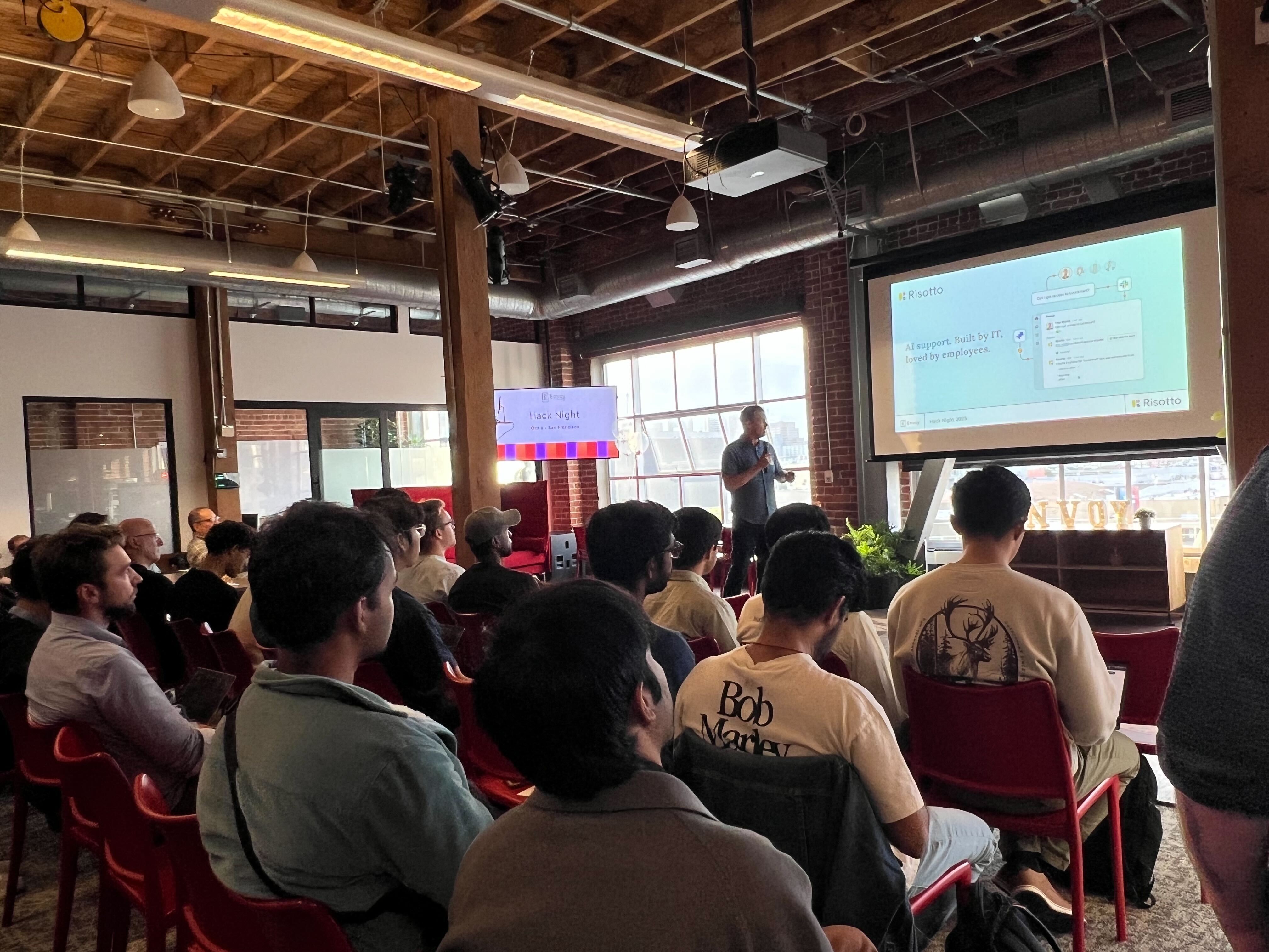 Audience seated facing a presenter speaking on AI support at an indoor event with slides and city view.