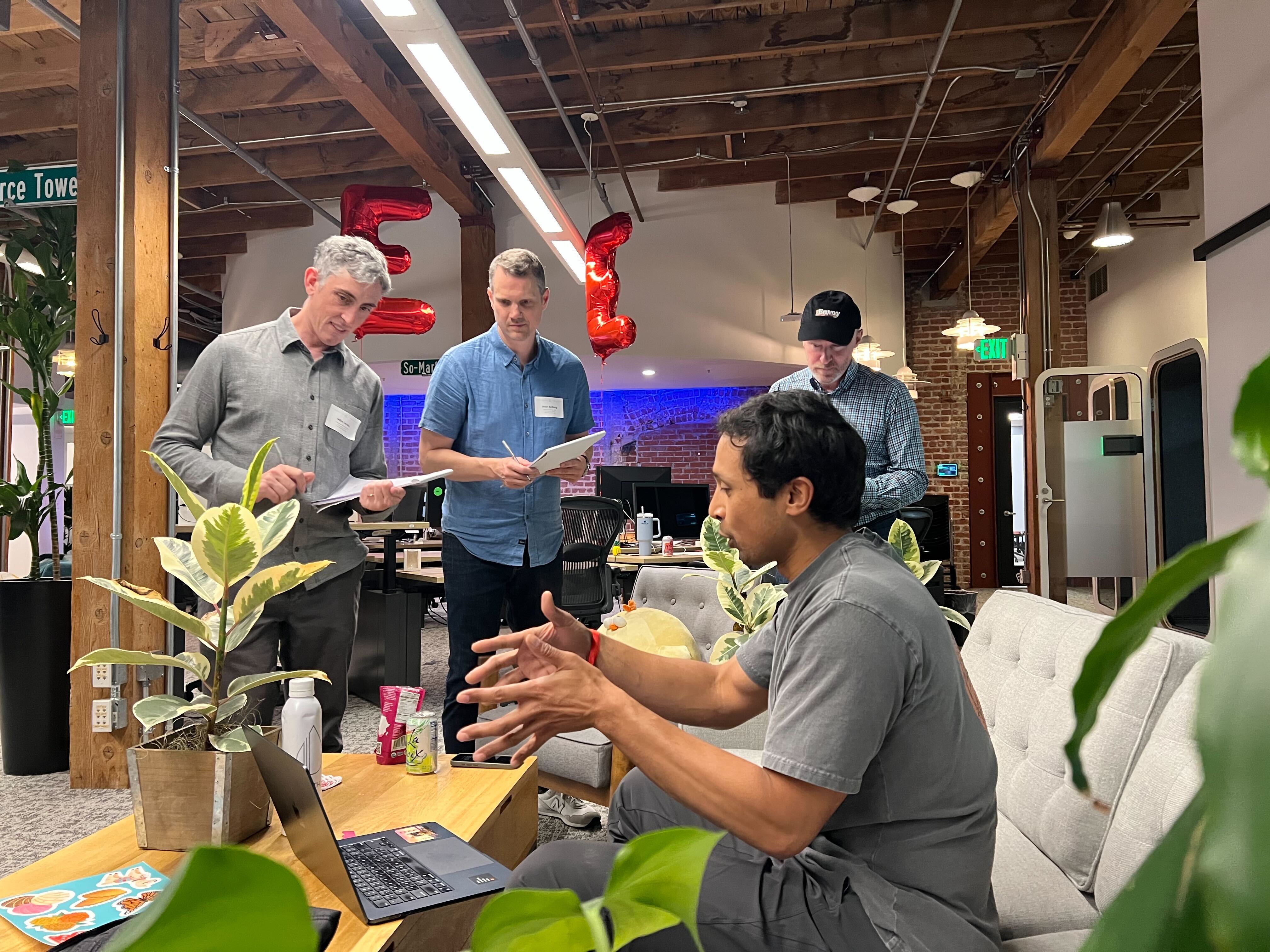 Man sitting on couch gestures while three men stand with clipboards in an office space with plants.