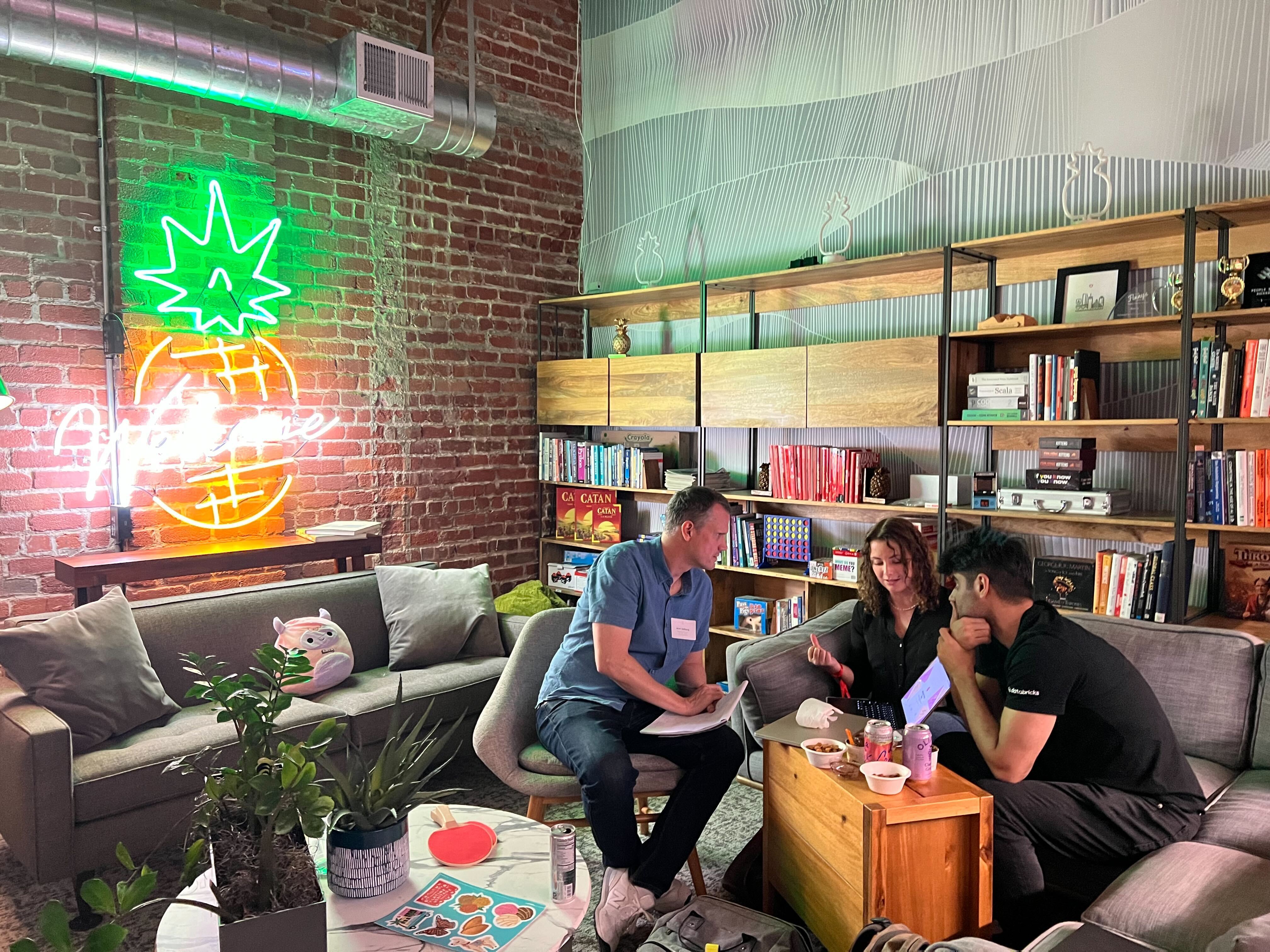 Three people sitting on sofas around a coffee table in a cozy room with bookshelves and a neon pineapple sign.