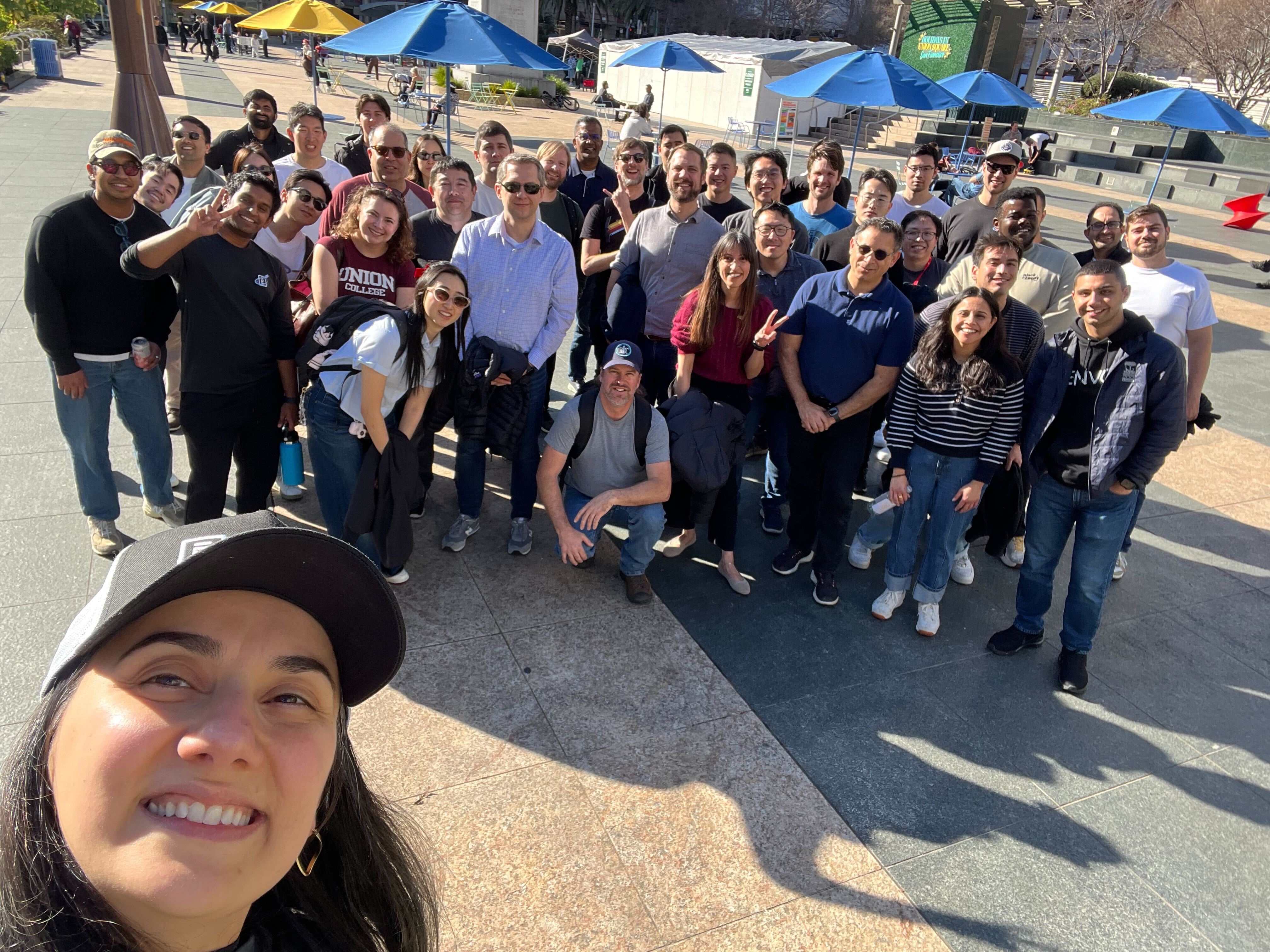 Large group of diverse adults smiling outdoors under blue umbrellas on a sunny day, one person taking a selfie.