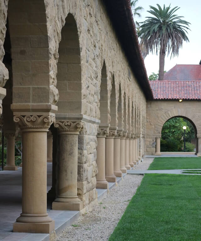 Stone archways with columns along a path next to a green lawn and a palm tree in the background.