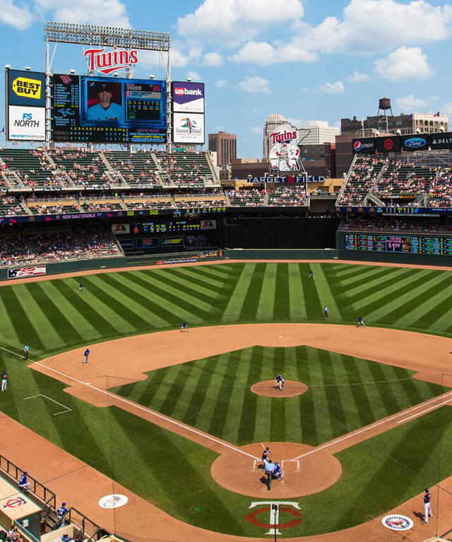 Baseball game at Minnesota Twins stadium with players on field and scoreboard under blue sky.