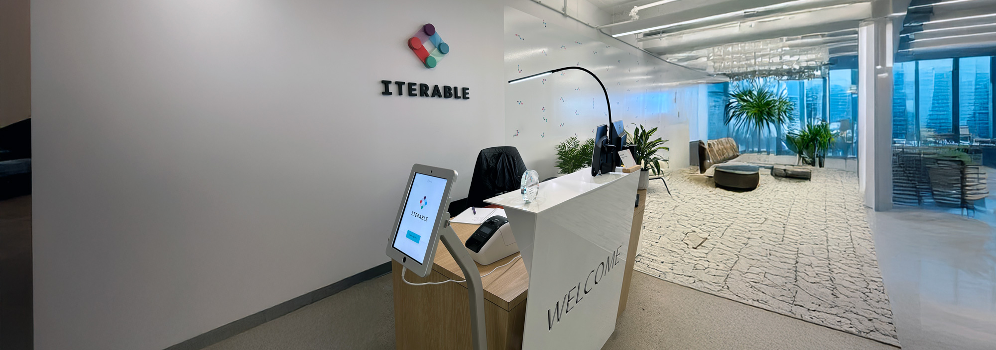 Modern office reception area with a white welcome desk, a tablet kiosk displaying the Iterable logo, and a wall-mounted Iterable logo on a plain wall; lounge area with potted plants, seating, and large windows with blue tinted glass in the background.
